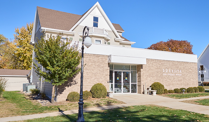 A two-story building with a mix of brick and siding, labeled BRANDTS 1884 PHARMACY MUSEUM, sits under a clear blue sky with trees and a sidewalk in front, not far from the historic Iles Brandt Chapel.