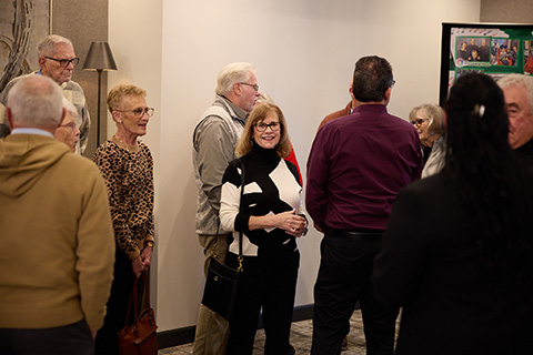 A group of older adults stands and converses in a room, possibly after discussing burial services. One woman in the center, wearing glasses and a black-and-white sweater, smiles at the camera while others are engaged in conversation.