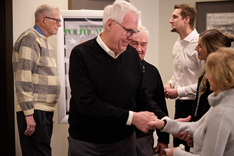 A group of people, mostly older men, stand indoors chatting and shaking hands. One man in a black sweater greets a woman in the foreground, while others discuss burial services nearby.