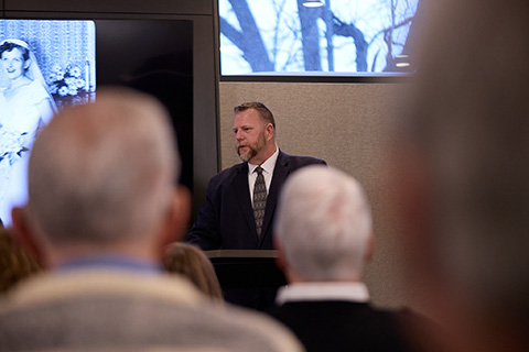 A man in a suit speaks at a podium in front of an audience during burial services. Behind him, a black-and-white photo of a bride is displayed on a screen, with some attendees' heads visible in the foreground.