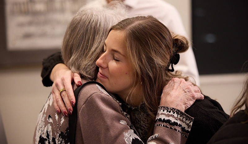 A young woman with light brown hair hugs an older woman with gray hair. Indoors, they share a warm embrace, finding comfort and support after discussing burial services together.