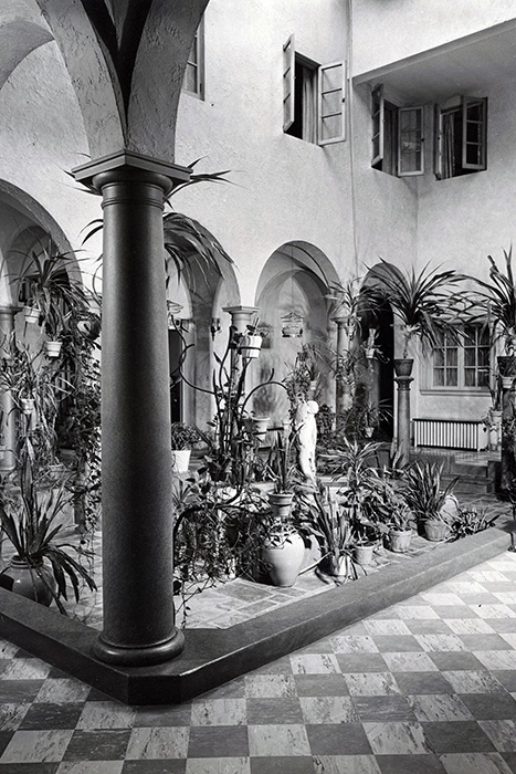 Black and white photo of Dunn's Chapel’s indoor courtyard with arched walkways, tiled floor, potted plants, and a small statue in the center. Open windows above let light pour in.