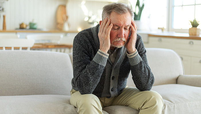 An older man sits on a couch with his elbows on his knees, holding his head in his hands and looking down, appearing stressed or deep in thought—perhaps grappling with grief when death occurs—in a bright, cozy living room.