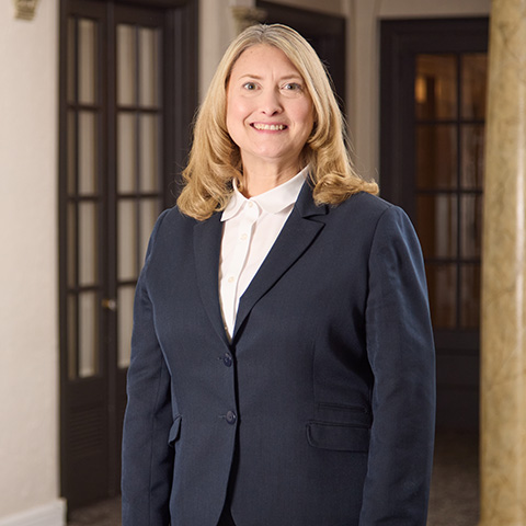 A woman with long blonde hair, wearing a navy blazer and white shirt, stands indoors in front of dark wooden doors and a marble column, smiling at the camera—reflecting the welcoming atmosphere created by caring staff.
