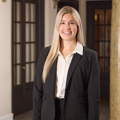 A woman with long blonde hair, wearing a black blazer and white blouse, stands indoors in front of tall wooden doors and a marble column, smiling warmly at the camera, reflecting the caring staff within.
