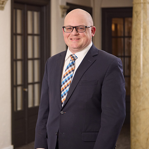 A man wearing glasses, a navy suit, and a colorful plaid tie stands smiling indoors near a marble column and wooden doors, embodying the warmth of the caring staff around him.