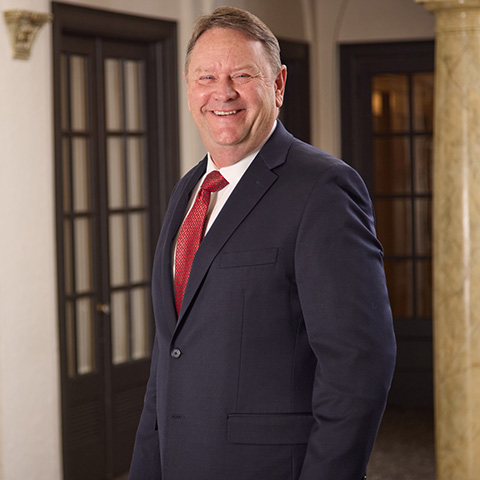 A man in a dark suit, white shirt, and red tie smiles while standing indoors near wooden doors and a marble column, reflecting the welcoming atmosphere created by the caring staff.