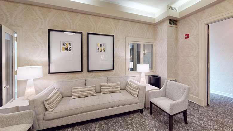 A modern living room with a beige sofa, three striped cushions, a matching armchair, two white table lamps on side tables, patterned wallpaper, framed art inspired by Dunn's Chapel, grey carpet, and an open doorway.