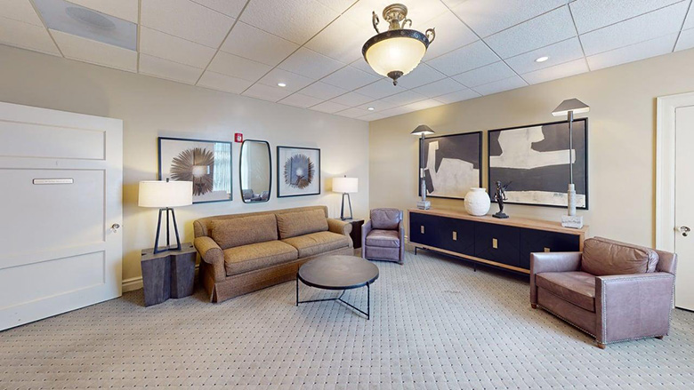 A modern lounge at Dunn's Chapel features a brown sofa, two leather armchairs, a round black coffee table, lamps, wall art, and neutral-colored carpeting beneath a ceiling light fixture.