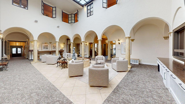 Spacious, elegant lounge with arched columns, a central fountain, neutral-toned seating, high ceilings, and a skylight. Upper windows overlook the room, creating an airy, Mediterranean-style atmosphere reminiscent of Dunn's Chapel.