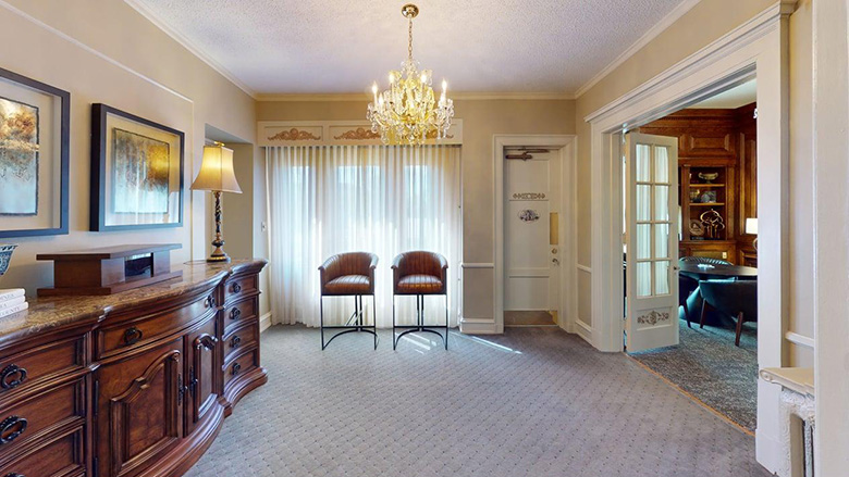 Elegant room with carpeted floor, wooden dresser, two chairs by a large curtained window, and chandelier overhead—evoking Dunn's Chapel style—with an open doorway revealing a wood-paneled office with desk and chair.