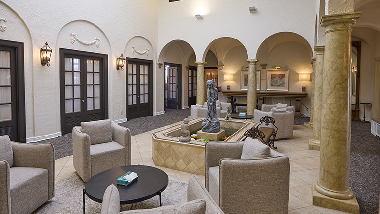 A spacious, elegant lounge at Dunn's Chapel features beige armchairs around a round table, a central fountain with a statue, arched doorways, decorative columns, and soft lighting creating a cozy, inviting atmosphere.