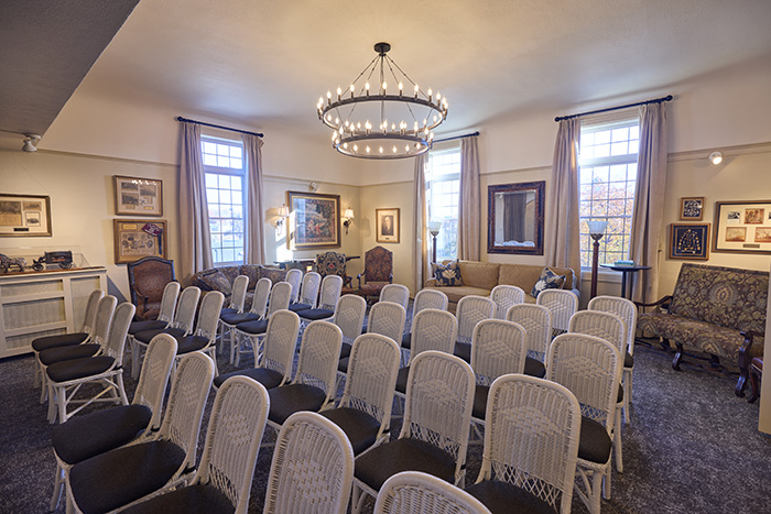 A well-lit room at Dunn's Chapel with rows of white wicker chairs facing forward, arranged for an event. The space features large windows, framed artwork on cream-colored walls, and a circular chandelier hanging from the ceiling.
