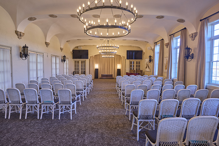 Spacious event room at Dunn's Chapel with rows of white wicker chairs facing a small stage, large chandeliers hanging from the ceiling, arched windows with curtains, and soft ambient lighting.