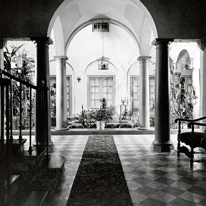 Black and white photo of Dunn's Chapel’s grand indoor courtyard with tall columns, arched ceilings, potted plants, hanging lanterns, and a patterned tile floor. Ornate furnishings are visible along the edges.