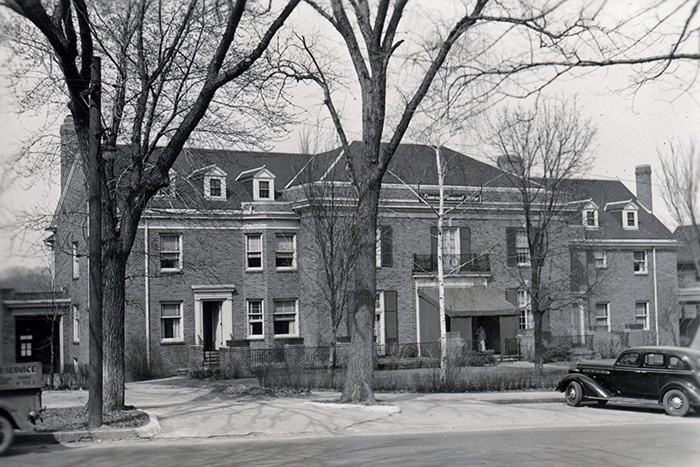 Black and white photo of Dunn's Chapel, a large brick building with multiple windows and dormers. Leafless trees stand in front, vintage cars line the street, capturing a scene from the early to mid-20th century.