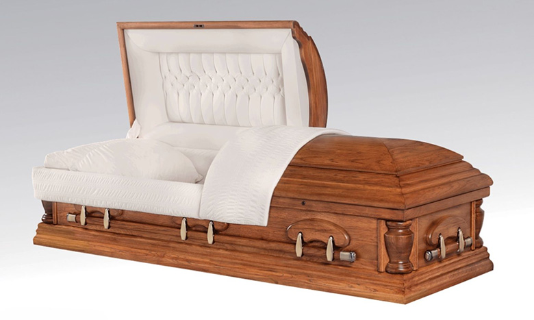 A piece of burial merchandise, this wooden casket with a white padded interior and partially open lid, draped in a white blanket, is displayed against a plain light gray background.