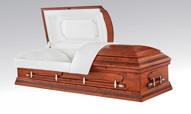 A wooden casket with detailed carvings is shown with its lid partially open, revealing a white padded interior and pillow. This elegant piece of burial merchandise rests against a plain, light background.