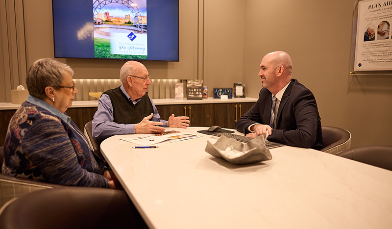 An elderly couple sits at a conference table talking with a professionally dressed man about cremation services. Pamphlets are on the table, and a large monitor with a presentation is visible in the background.
