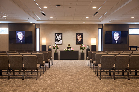 A modern funeral chapel with rows of empty chairs facing an altar adorned with flowers, candles, and framed photos of a smiling man. Two large screens display his portrait, creating a peaceful setting for thoughtful reflection and cremation services.