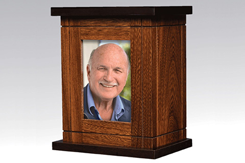 A wooden cremation urn with a rectangular shape, featuring a framed photograph of an older man with a bald head and mustache, smiling, set into the front panel—a dignified tribute often chosen for cremation services.