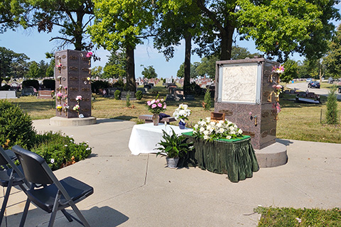 A memorial service setup in a cemetery features a table covered in flowers and a green cloth, surrounded by chairs. Two mausoleum structures adorned with floral arrangements suggest options for traditional burials or cremation services. Trees and gravestones line the background.