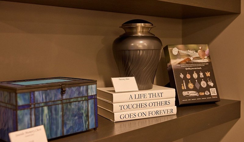 A shelf displays a decorative urn on stacked books titled A Life That Touches Others Goes on Forever, a blue stained glass box, and a catalog featuring cremation packages and memorial jewelry options.