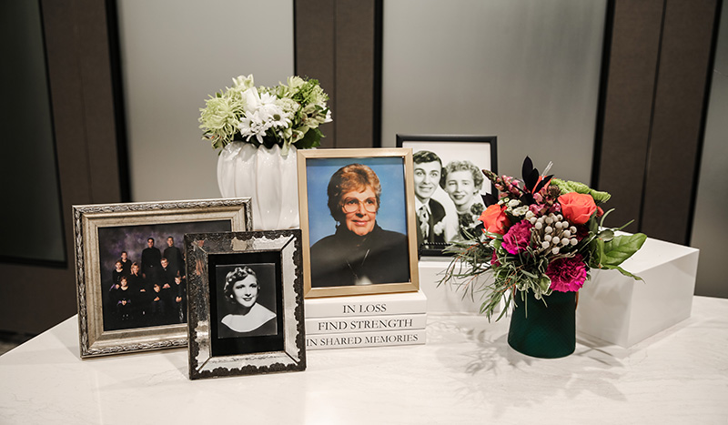 A table displays framed family photos, a white vase with white flowers, a green vase with colorful flowers, a white box, and books titled IN LOSS, FIND STRENGTH, and IN SHARED MEMORIES—reflecting the comfort offered by thoughtful cremation packages.