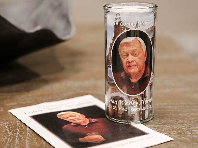 A memorial candle and a printed card are displayed on a table, both featuring a photo of an elderly man with white hair. Often included in cremation packages, the candle reads “In Loving Memory” with his name and dates of birth and death.