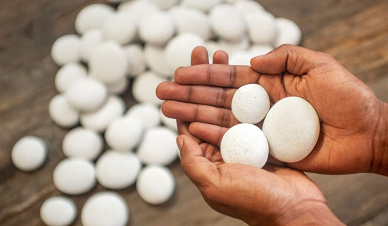 A pair of hands holds several smooth, white stones of different sizes, often used as cremation merchandise, while many more similar stones are scattered on a wooden surface in the background.