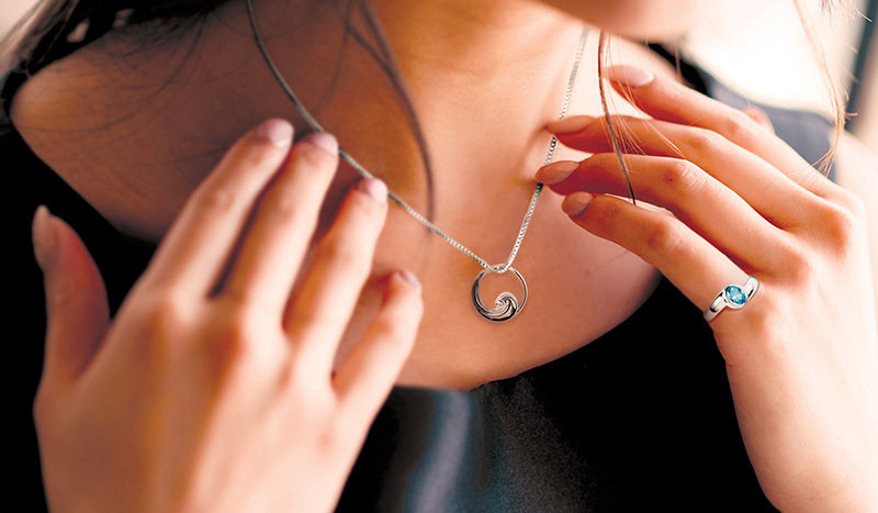 A woman wearing a black top touches a silver cremation merchandise necklace with a circular pendant around her neck. She also wears a silver ring with a blue gemstone on her right hand.