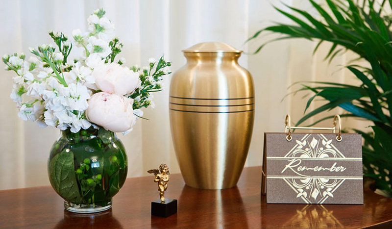 A gold cremation merchandise urn sits on a wooden table, flanked by a vase of white and pink flowers, a small angel figurine, and a standing sign that reads Remember. A green plant is in the background.