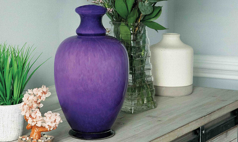 A large purple urn, part of elegant cremation merchandise, sits on a wooden cabinet beside a small potted plant, a white vase, a clear glass vase with green leaves, and artificial pink flowers.