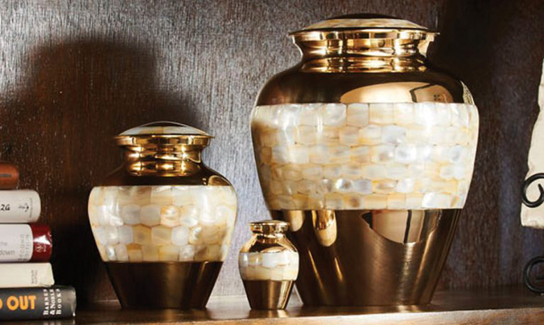 Three decorative urns of varying sizes, designed as elegant cremation merchandise with gold bases and lids, feature a band of pearly, mosaic-like tiles and are displayed on a wooden shelf beside a small stack of books.