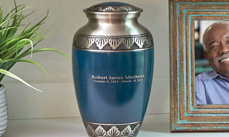 A blue and silver cremation merchandise urn engraved for Robert James Matthews, dated October 2, 1942 – March 18, 2019, sits on a shelf beside a green plant and part of a framed photo of a smiling older man.