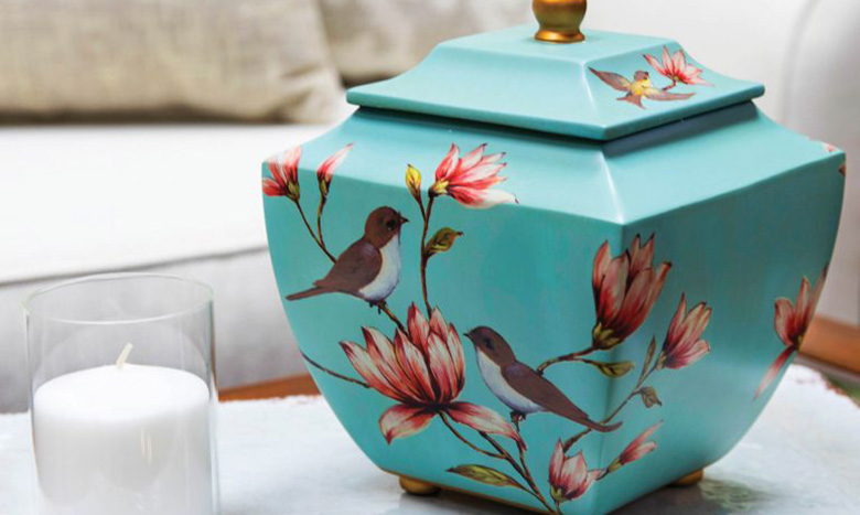 A decorative teal ceramic jar with a lid, featuring painted birds and pink flowers, sits next to a white candle in a glass holder on a table—an elegant addition to your cremation merchandise selection.