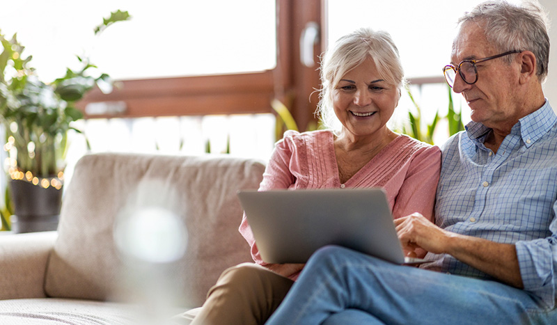 An older couple sits together on a sofa, smiling as they look at a laptop. The bright room, filled with green plants, sets a calm atmosphere while they explore funeral pre-planning options online.