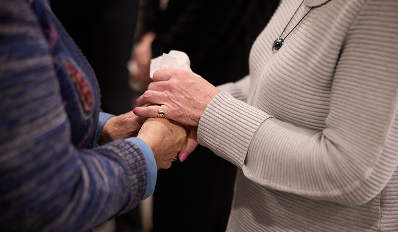 Two people stand facing each other, holding hands in a comforting gesture. One, perhaps discussing funeral pre-planning, wears a gray sweater and necklace; the other, in a patterned blue sweater, holds a tissue.