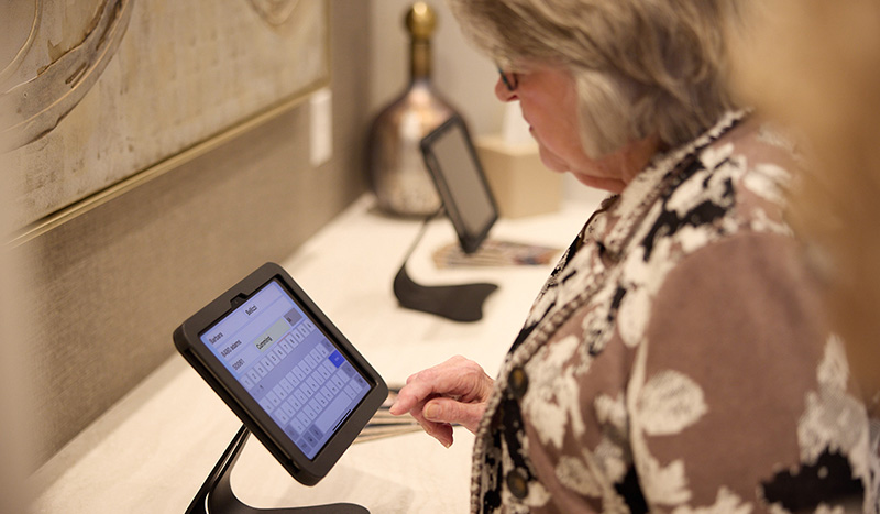 An older woman uses a touchscreen tablet mounted on a stand, typing on a virtual keyboard in what appears to be a public setting, possibly for funeral pre-planning registration or information.