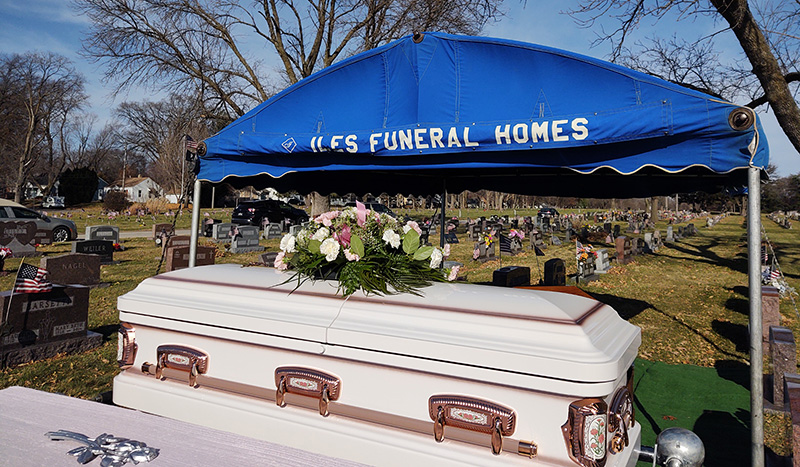 A white casket with pink and white flowers rests under a blue canopy labeled ILES FUNERAL HOMES at a cemetery, with gravestones and trees beyond—a peaceful setting that highlights the importance of funeral pre-planning.
