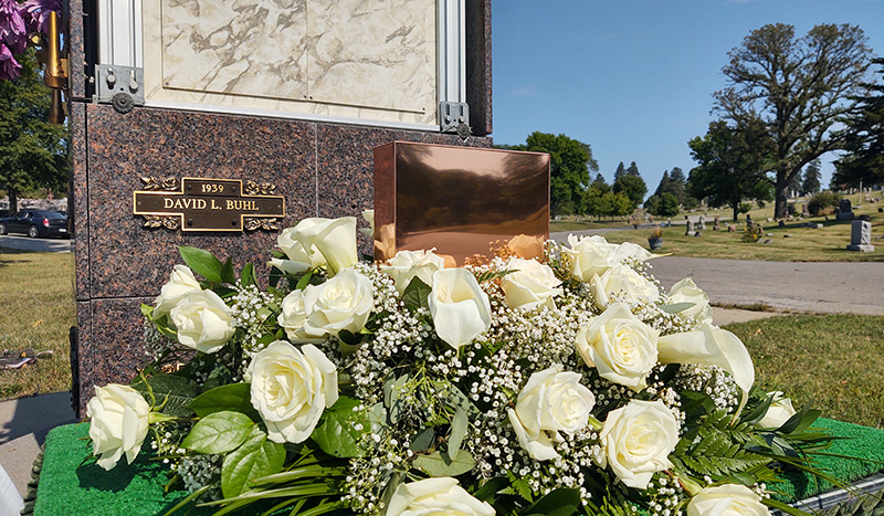 A bronze urn and a bouquet of white roses rest in front of a mausoleum niche labeled “David L. Buhl 1939.” The peaceful cemetery scene highlights the importance of funeral pre-planning for honoring loved ones.