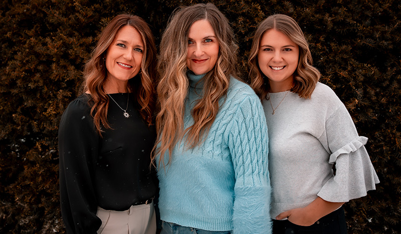 Three women standing close together outdoors, smiling at the camera. They have wavy hair and are wearing casual sweaters and blouses, sharing a moment of connection—perhaps discussing important topics like funeral pre-planning—against a dark, leafy backdrop.