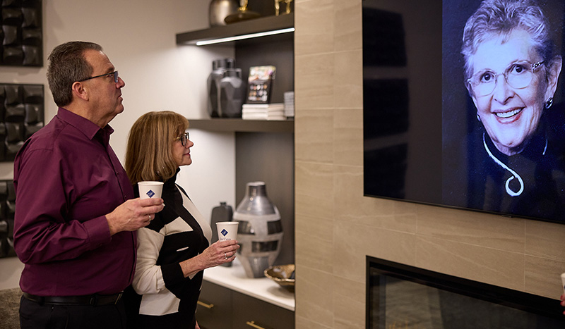 A man and woman holding coffee cups stand together, looking at a large portrait of an older woman smiling—a tribute to Iles Funeral Homes History—on a wall inside a modern, warmly lit room with decorative shelves.