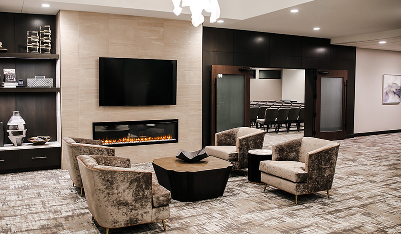Modern lounge area with four plush chairs around a wooden coffee table, a wall-mounted TV above a linear fireplace, built-in shelves on the left, and a glass door leading to Central Iowa Funeral Care service seating.