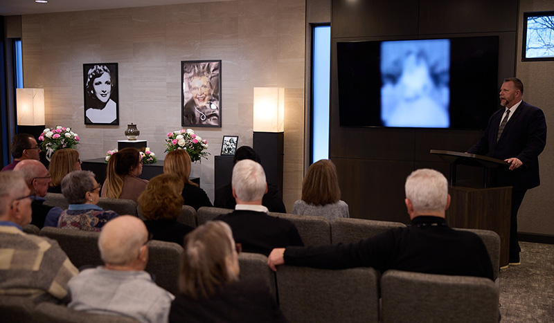 A group of people sit in a room facing a speaker at a podium. Behind him, a blurry photo is displayed on a screen. Two portraits and flower arrangements suggest a memorial or funeral service by Central Iowa Funeral Care.