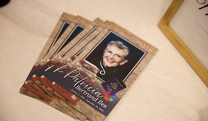 A stack of memorial cards from Central Iowa Funeral Care features a photograph of an older woman wearing glasses and smiling. The cards display the name “Patricia Bertrand Illes,” her birth and death dates, and decorative elements.