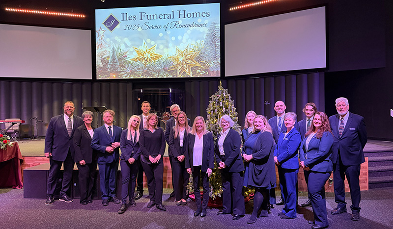 A group of people in formal attire stand and smile in front of a decorated Christmas tree on a stage. Behind them, a large screen displays Iles Funeral Homes 2023 Service of Remembrance, honoring Central Iowa Funeral Care traditions.