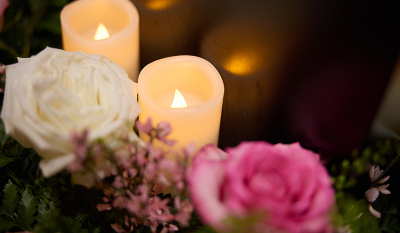 Two lit candles are surrounded by a white rose, a pink rose, and small pink flowers, creating a warm and peaceful ambiance—reflecting the comforting support of Central Iowa Funeral Care.