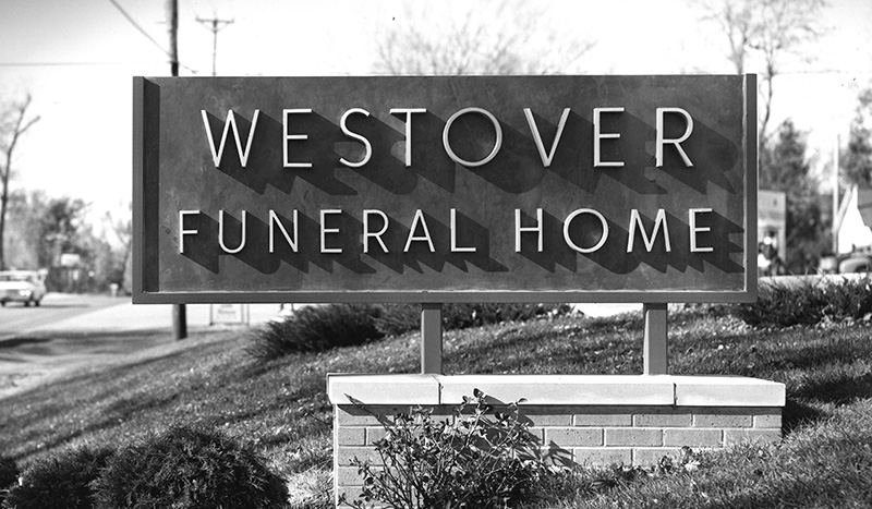 A large outdoor sign reads “Westover Funeral Home” in bold letters, set on a brick base with bushes and grass in the foreground—a classic scene reflecting Iles Funeral Homes History, shown here in black and white with a street, trees, and cars beyond.