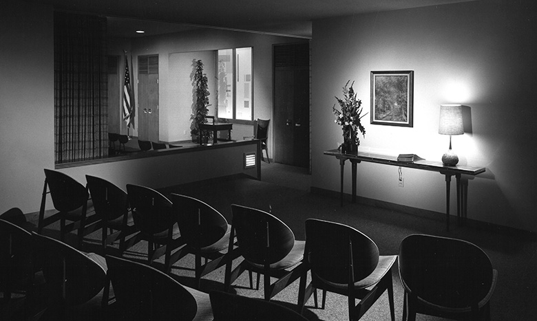 Black-and-white photo of a small, empty courtroom with rows of chairs, a table with a lamp and flowers, an American flag in the background, and a framed picture on the wall.